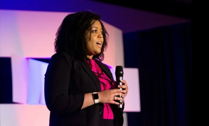 An African American woman speaks into a microphone while standing on a stage.